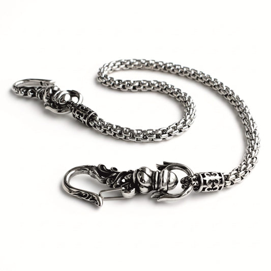 Geflochtene Edelstahl-Hosenkette mit verzierten Ornamenthaken, leicht geschwärztem Finish und schlangenartiger Struktur auf hellem Hintergrund.